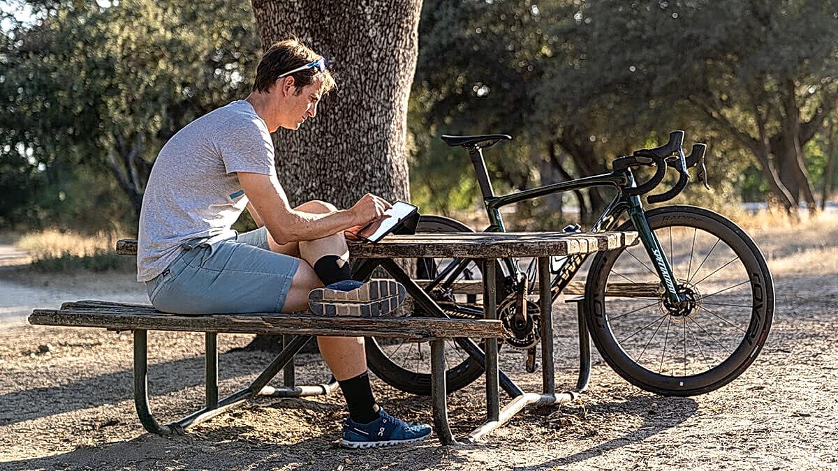 Running Cycling Coach Sitting On Laptop At The Park On Picnic Table Next To Back Setting Prices For Coaching Services